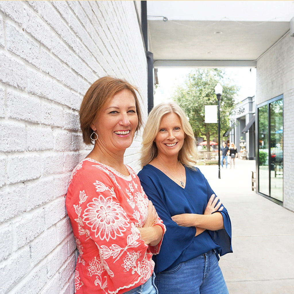 The founders of Tiny Type Studios and creators of Adoptionly Yours cards, Jayne and Stacy, leaning against a white brick wall, arms crossed and smiling.