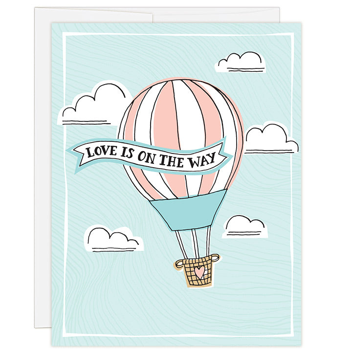 4.25 x 5.5 inch adoption waiting card. Blank inside. Simple and charming illustration style. Title Love Is On The Way. Drawing of a pink stripped hot air balloon with a brown woven basket rising high up in the sky with white fluffy clouds. Title is in a ribbon draped across the hot air balloon.
