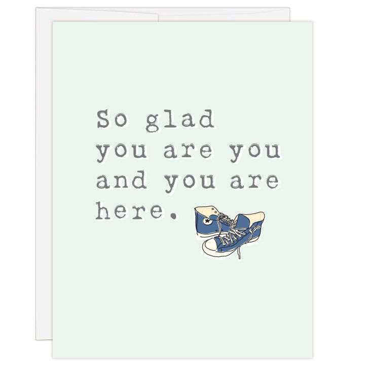 4.25 x 5.5 inch greeting card. Blank inside. Cover is pale green with gray lettering and colored line illustration of a pair of blue high-top tennis shoes. Typewriter text reads: So glad you are you and you are here.