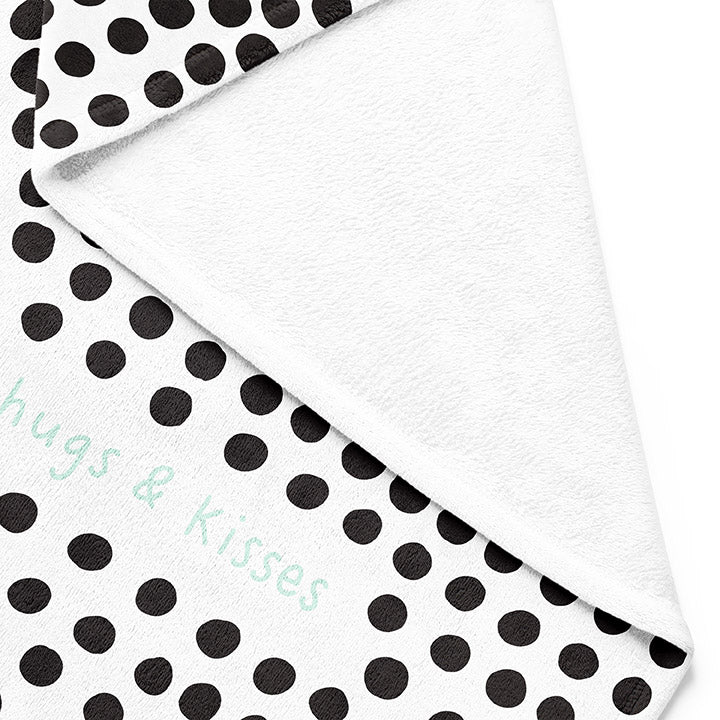 Backside of white 30 by 40 inch silky-soft fleece blanket that is hypoallergenic. Perfect size for a toddler or child. Bright white blanket printed on one side only. 3 inch hand drawn black dots scattered across blanket with the words hugs & kisses hand drawn in a mint green color. A small Adoptionly Yours heart logo by itself in mint green in lower left corner of blanket on front. 