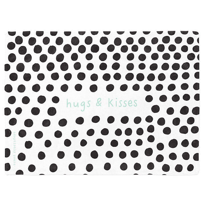 30 by 40 inch silky-soft fleece blanket that is hypoallergenic. Perfect size for a toddler or child. Bright white blanket printed on one side only. 4 inch hand drawn black dots scattered across blanket with the words hugs & kisses hand drawn in a mint green color. The words adoptionly yours in mint green typewriter font on lower left corner of blanket on front. 