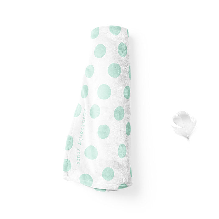 Rolled up 50 by 60 inch silky-soft fleece blanket that is hypoallergenic. Perfect size for a toddler or child. Bright white blanket printed on one side only. 4 inch hand drawn dots scattered across blanket in mint green with the words hugs & kisses hand drawn in a peach color. The words adoptionly yours in mint green typewriter font on lower left corner of blanket on front. 