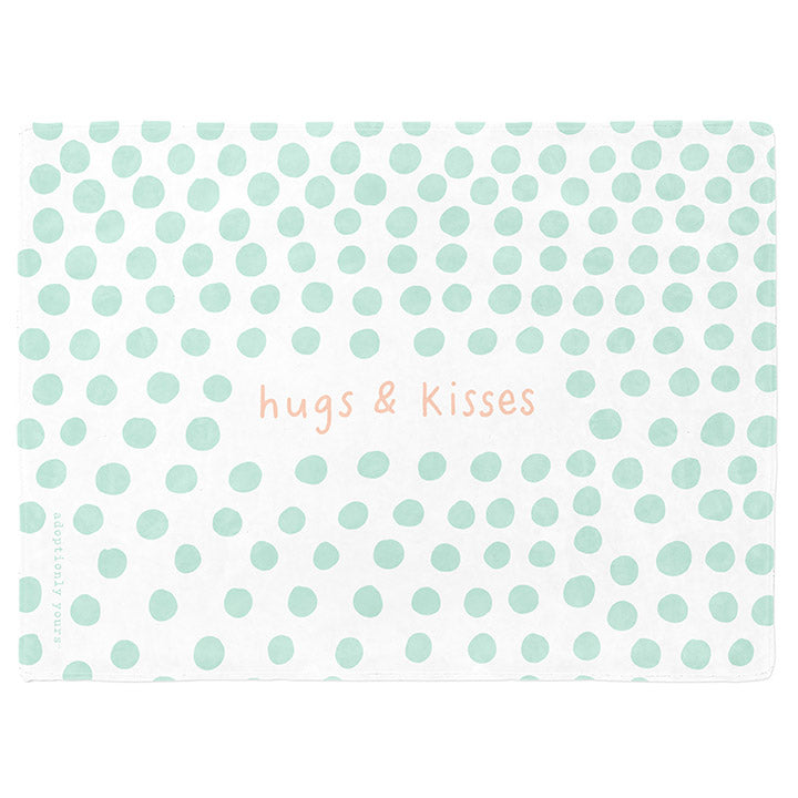 50 by 60 inch silky-soft fleece blanket that is hypoallergenic. Perfect size for a toddler or child. Bright white blanket printed on one side only. 4 inch hand drawn dots scattered across blanket in mint green with the words hugs & kisses hand drawn in a peach color. The words adoptionly yours in mint green typewriter font on lower left corner of blanket on front. 