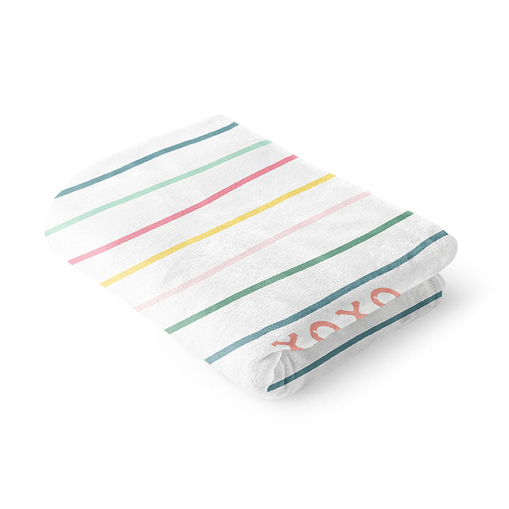 Folded 50 by 60 inch silky-soft fleece blanket that is hypoallergenic. Perfect size for an adult. Bright white blanket printed on one side only. 13 colorful hand drawn stripes of pink, green, blue, light teal, magenta and bright yellow run horizontally with the words xoxo, always in typewriter font. Adoptionly Yours heart logo by itself in pink in lower left corner of blanket on front. 