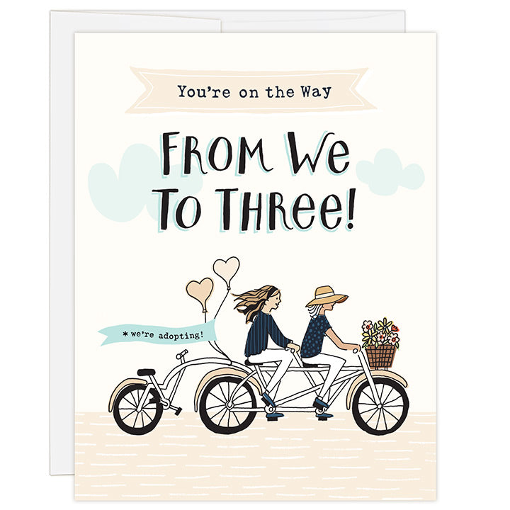 4.25 x 5.5 inch adoption greeting card. Blank inside. Simple and charming illustration style. Title You’re on the Way From We to Three!  Main image is two women on a tandem bicycle. The woman in front is wearing large brim hat and the woman in back is wearing a head band and has long hair. The third wheel of the bike has a back seat and it is empty. There are flowers in the basket and balloons on the back of the bike with the words we’re adopting!
