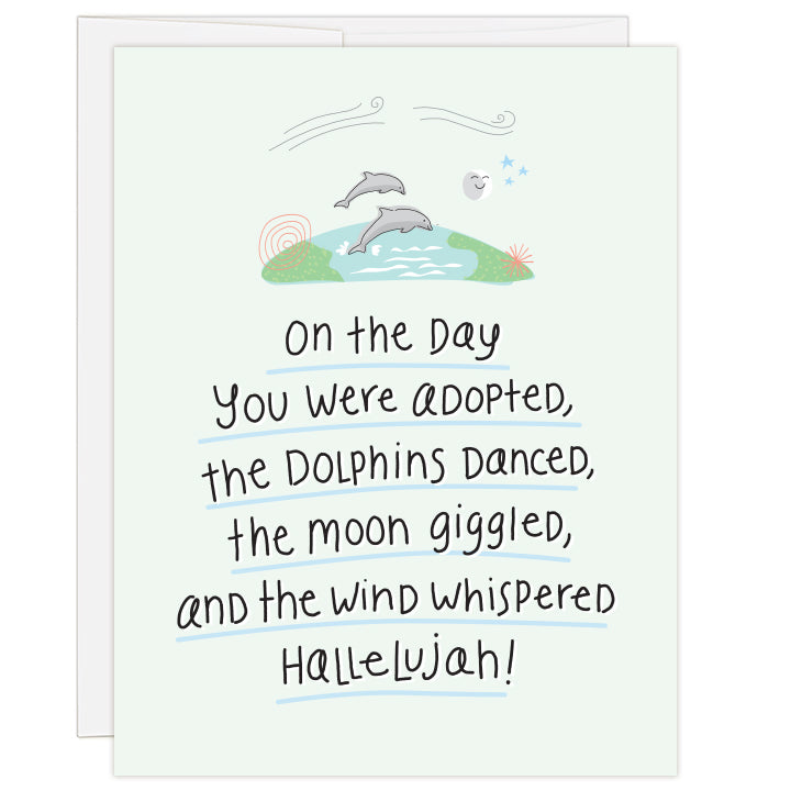 4.25 x 5.5 inch greeting card. Blank inside. Pale green cover features a storybook illustration of jumping dolphins above the earth, with a smiling moon and stars and wisps of wind. Large hand-illustrated text reads: On the day you were adopted, the dolphins danced, the moon giggled, and the wind whispered hallelujah!