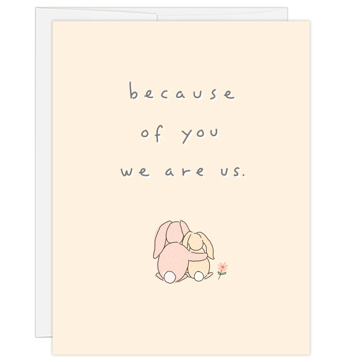 4.25 x 5.5 inch greeting card. Blank inside.  Simple and charming line illustration of one bunny with arm around smaller bunny, as seen from behind, with small flower to the side. On pale peach background. Text: because of you, we are us.