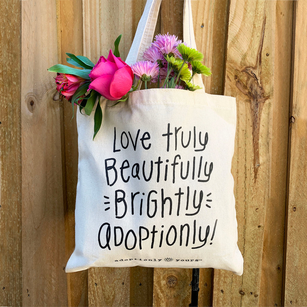 Photo of adoptionly yours natural cotton canvas tote filled with brightly colored flowers. Tote reads: love truly, beautifully, brightly, adoptionly. 